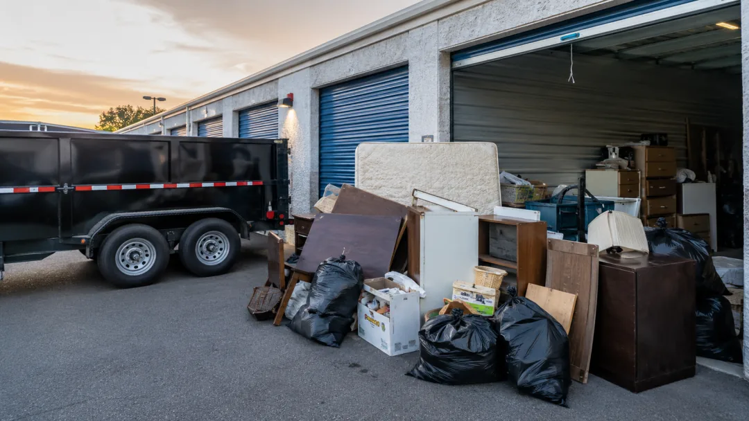 Dump trailer parked outside a storage unit during a cleanout, furniture and bags alongside
