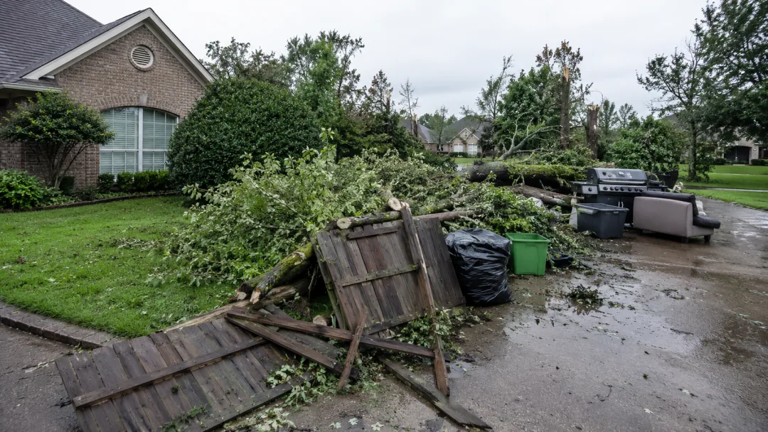 Downed tree limbs and broken fence boards scattered across a wet lawn after a storm