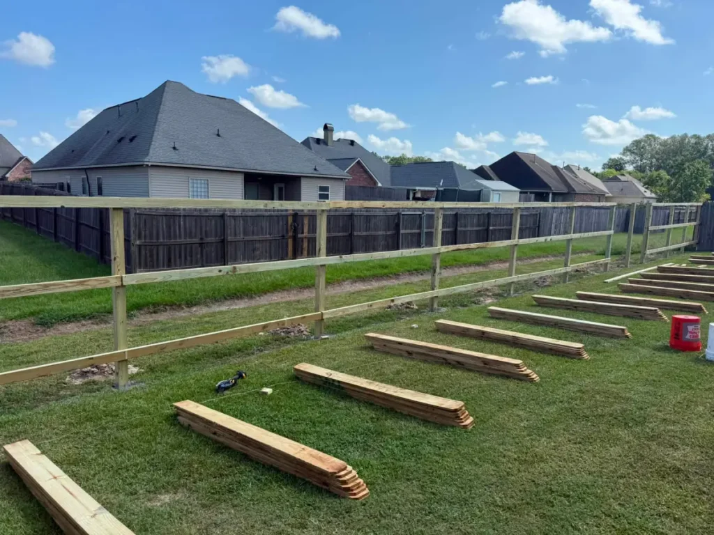 A fence installation in progress in Denham Springs: posts and rails set, with fresh pickets laid out on the grass ready to hang.