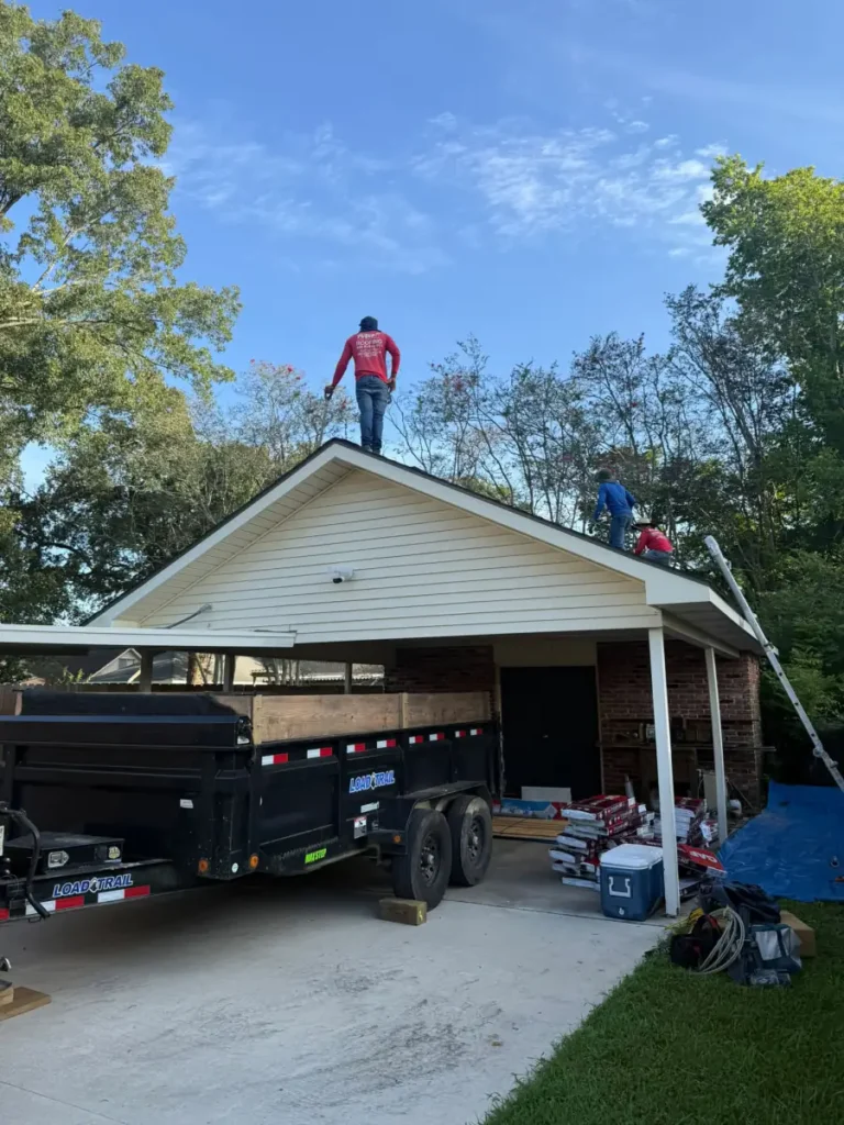 Crew working on a roof tear-off with an Acadian Services dump trailer staged in the carport driveway to catch the debris.