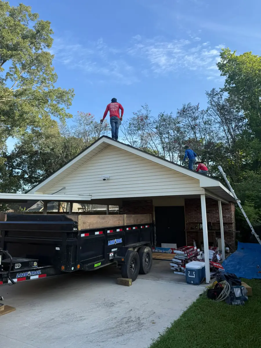 Crew working on a roof tear-off with an Acadian Services dump trailer staged in the carport driveway to catch the debris.