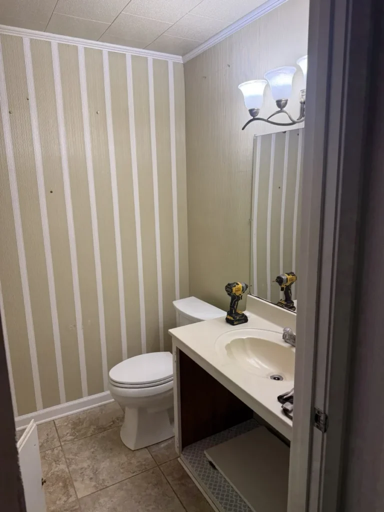 A half bathroom before demolition, with striped wallpaper, a toilet, and a cultured marble vanity, and a drill set on the counter ready to start.